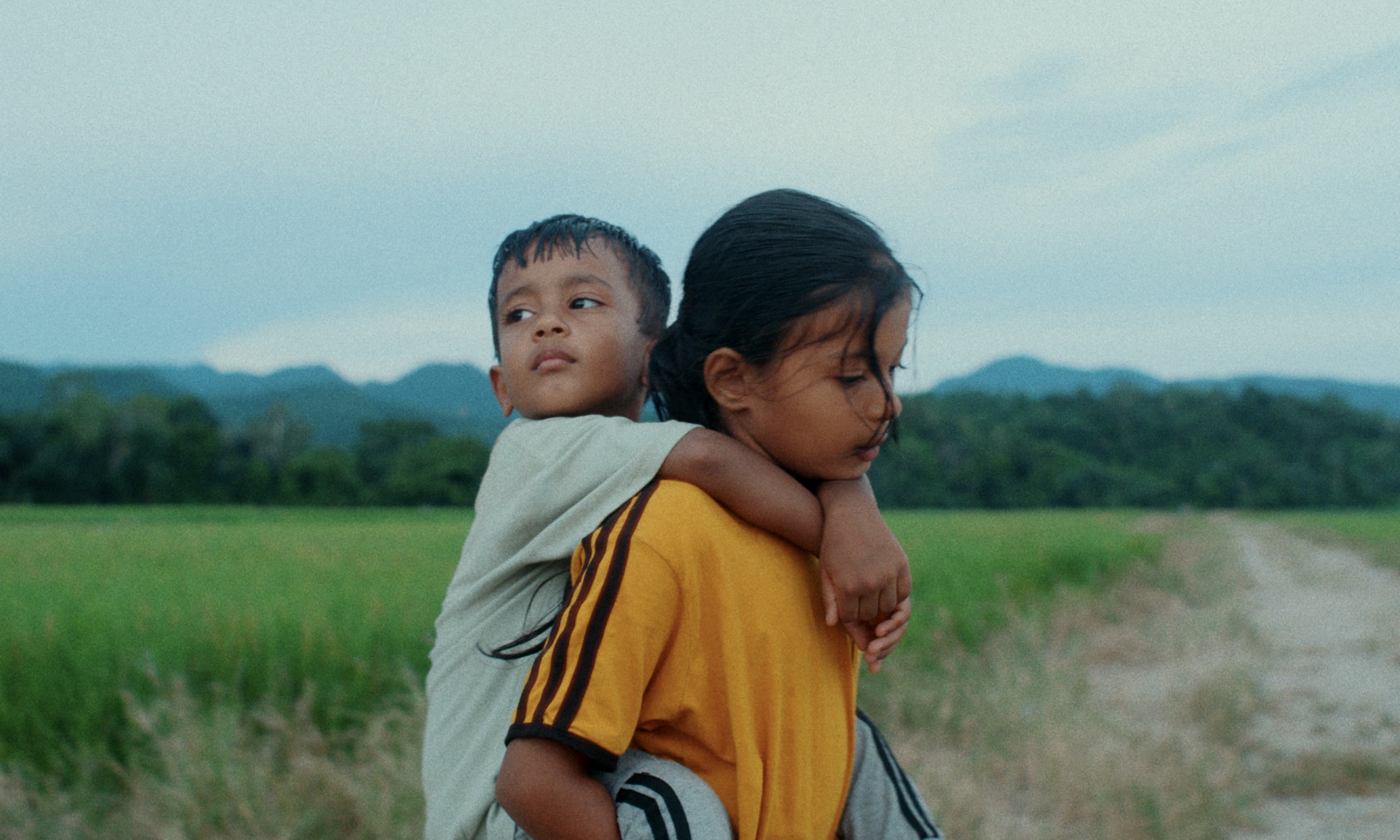 Dans « Les Fleurs du Manguier », Akio Fujimoto filme le périple de deux jeunes Rohingyas, Shafi et Somira, interprétés par Shofik Rias Uddin et Shomira Rias Uddin. Sur l'image la petite fille porte le petit garçon sur son dos au milieu d'un champ.