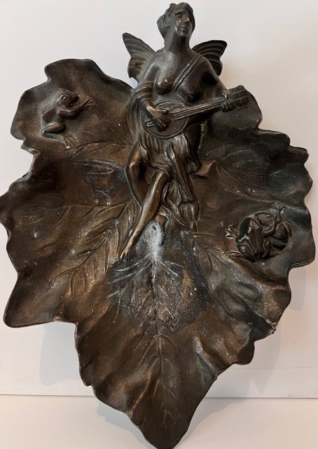 Une sculpture murale en métal représentant une femme ailée jouant d’un instrument sur une grande feuille décorative.
