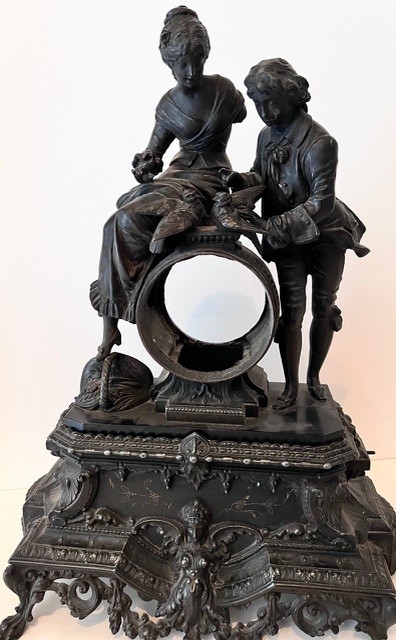 Une sculpture décorative sombre représentant deux personnages en tenue ancienne autour d’un support circulaire ornementé.