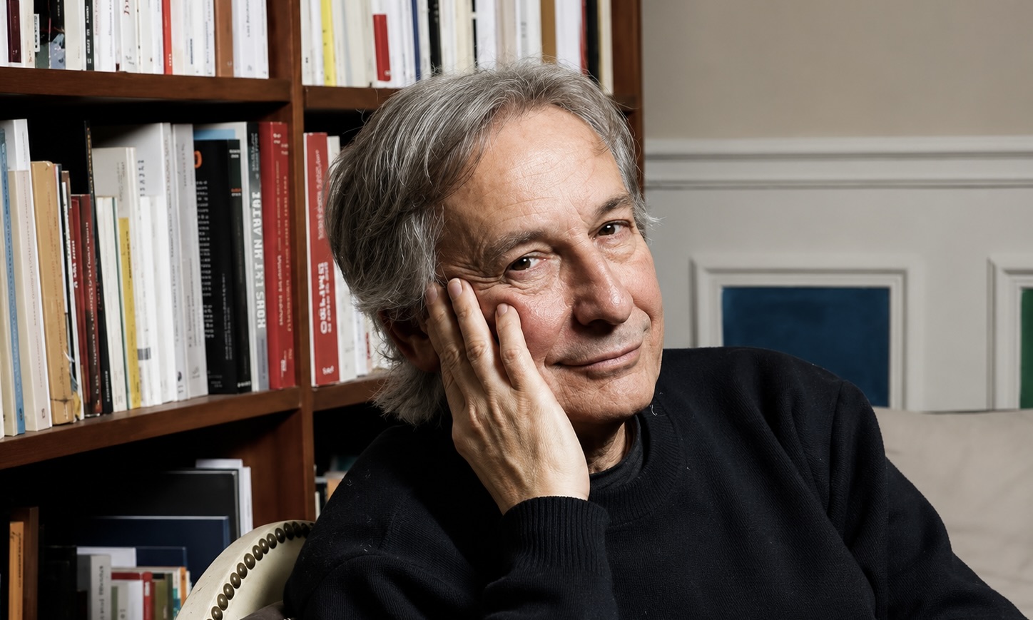L’écrivain Jean Rouaud pose assis devant bibliothèque.