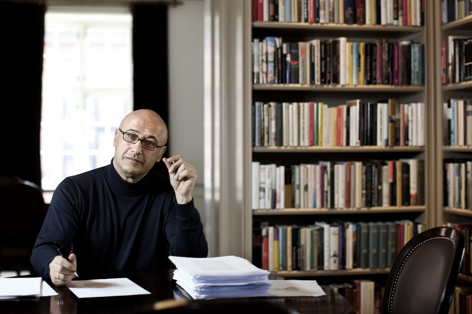 Gabi Gleichmann dans son bureau, il est assis en train d'écrire, on voit une bibliothèque à droite.