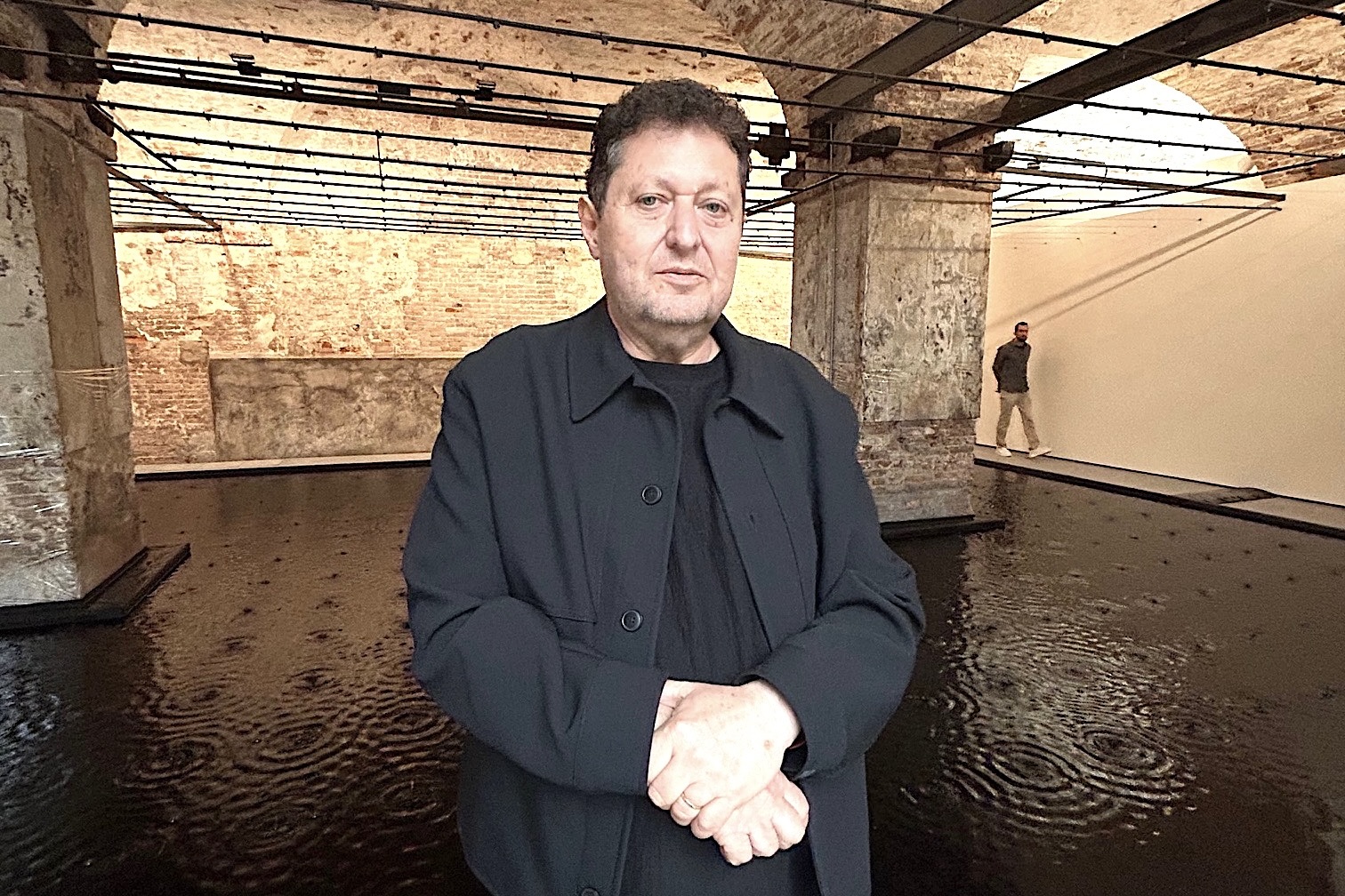 À Biennale, Belu-Simion Fainaru pose devant son œuvre « Rose of Nothingness », au Pavillon d’Israël. Derrière lui, on voit un bassin avec de l'eau, le fond du bassin est noir, il y a deux piliers en pierres et des murs en pierres. Le bassin est en intérieur.