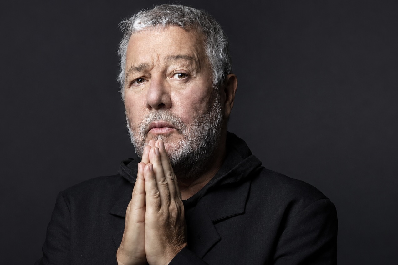 Le designer et architecte français Philippe Starck pose devant un fond gris, il a les mains jointes sous le menton.