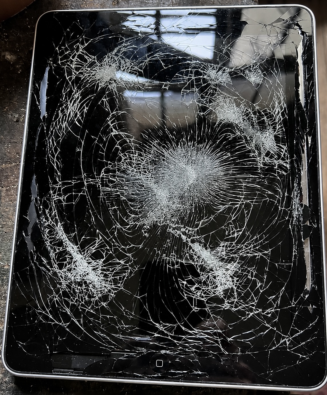Une tablette tactile à l’écran entièrement fissuré est photographiée à plat sur une surface sombre.