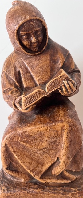Statuette en bois représentant une personne encapuchonnée assise, lisant un livre ouvert.
