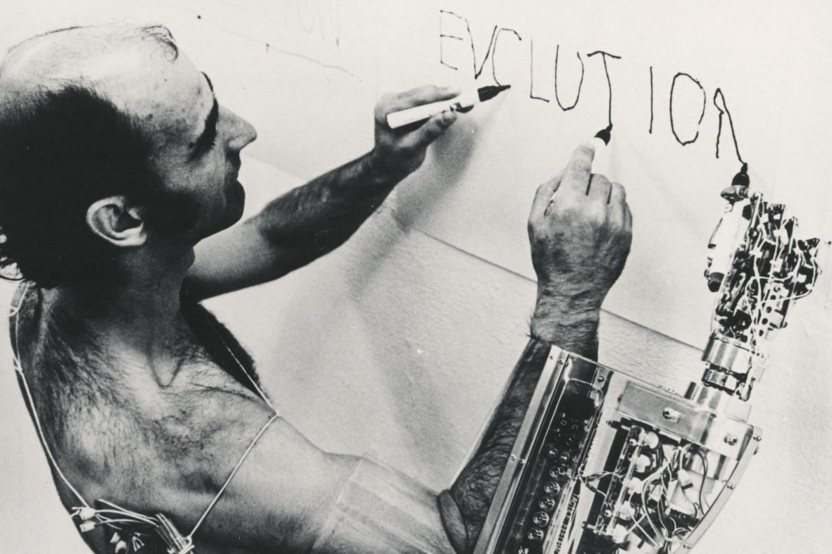 En 1982, à Tokyo, l’artiste Stelarc écrit sur un mur à l’aide d’un bras mécanique relié à son corps, explorant la fusion de l’homme et de la machine.